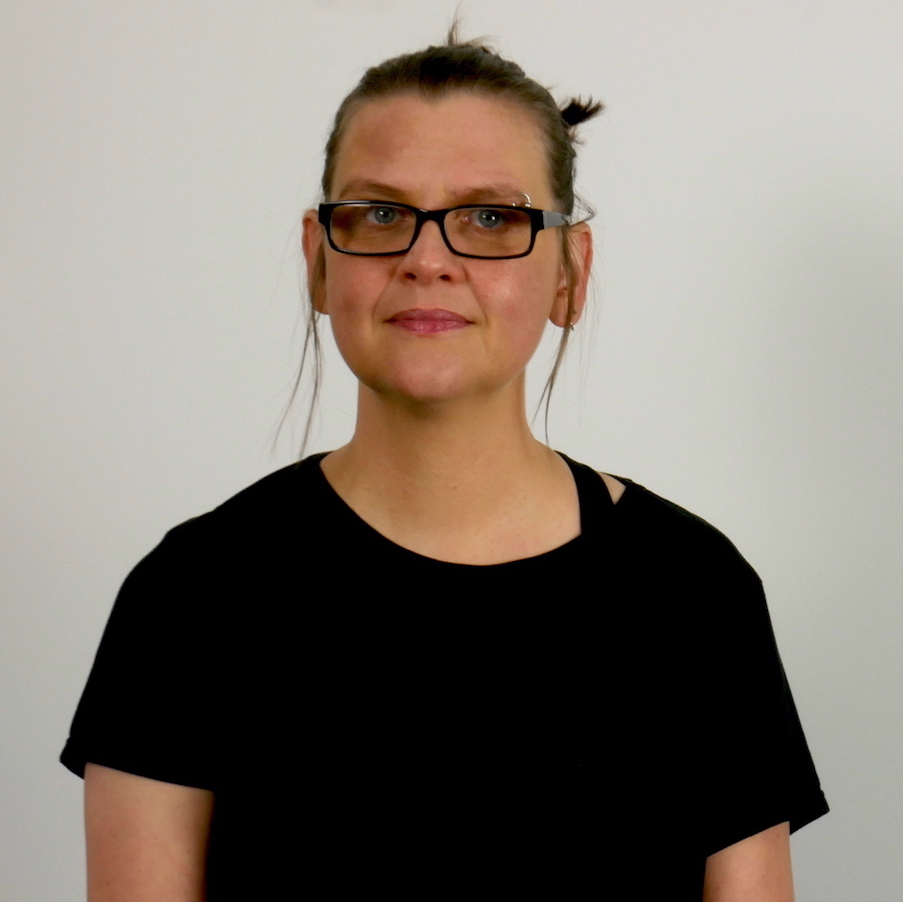 A portrait of me, by me, in a black t-shirt and black glasses. I'm a white person in my 40s with a silver hoop eyebrow piercing and melasma on my forehead. My shoulder-length brown hair is up in a messy bun with some wisps hanging down. I look slightly to the side of the camera with an almost-smile. The vibe is something like "I have a queer crip secret".