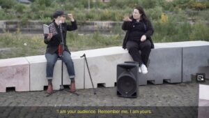 An outdoor reading on a ramped concrete sculpture in front of grass and concrete graving docks. Left is San Alland, wearing jeans, a black shirt and jacket, red boots and a grey cap, holding a copy of the book Stairs and Whispers. San is seated on the ramp in front of a mic. To their right is Bea Webster, seated a metre away further up the ramp. Bea is using BSL and is behind a small amplifier. San and Bea look at each other. San is white and Bea is Scottish-Thai with long dark hair. A red polka dot facemask hangs from the mic stand. Captions read "I am your audience. Remember me, I am yours."