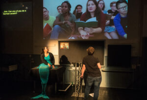 A live theatre performance. On the far left, captions are projected onto the wall. The say "Shit. Can any of you do a German accent?" To the right of the captions is a large video projection of nine people holding colourful chapbooks. A Black woman is speaking. An onscreen caption reads: "You are in an elevator." Below the screen and looking up at it is a Chineses BSL interpreter. She has long hair, is dressed like a mermaid and sits on a table. To her right is San, who has their back to the camera as they talk to the screen. They have short brown hair and lean on a black cane.